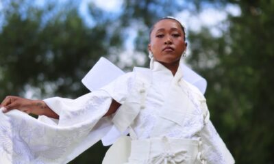 Naomi Osaka holding out the wide sleeve of her embroidered white gown during an outdoor fashion shoot.