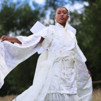Naomi Osaka holding out the wide sleeve of her embroidered white gown during an outdoor fashion shoot.