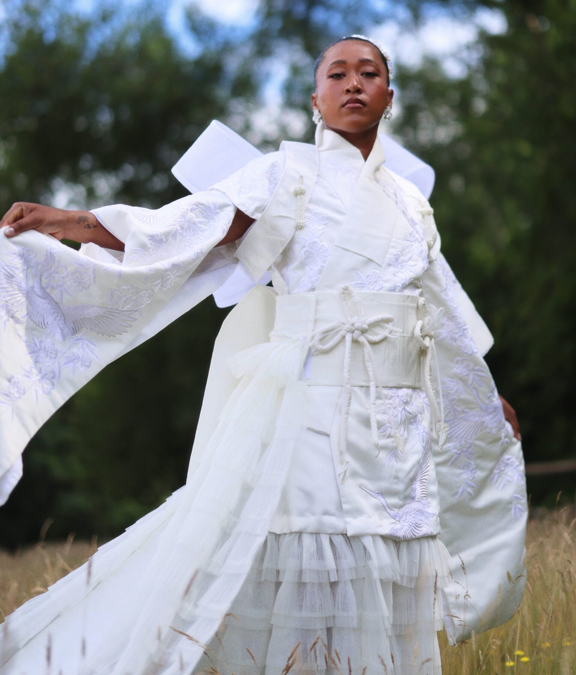 Naomi Osaka holding out the wide sleeve of her embroidered white gown during an outdoor fashion shoot.