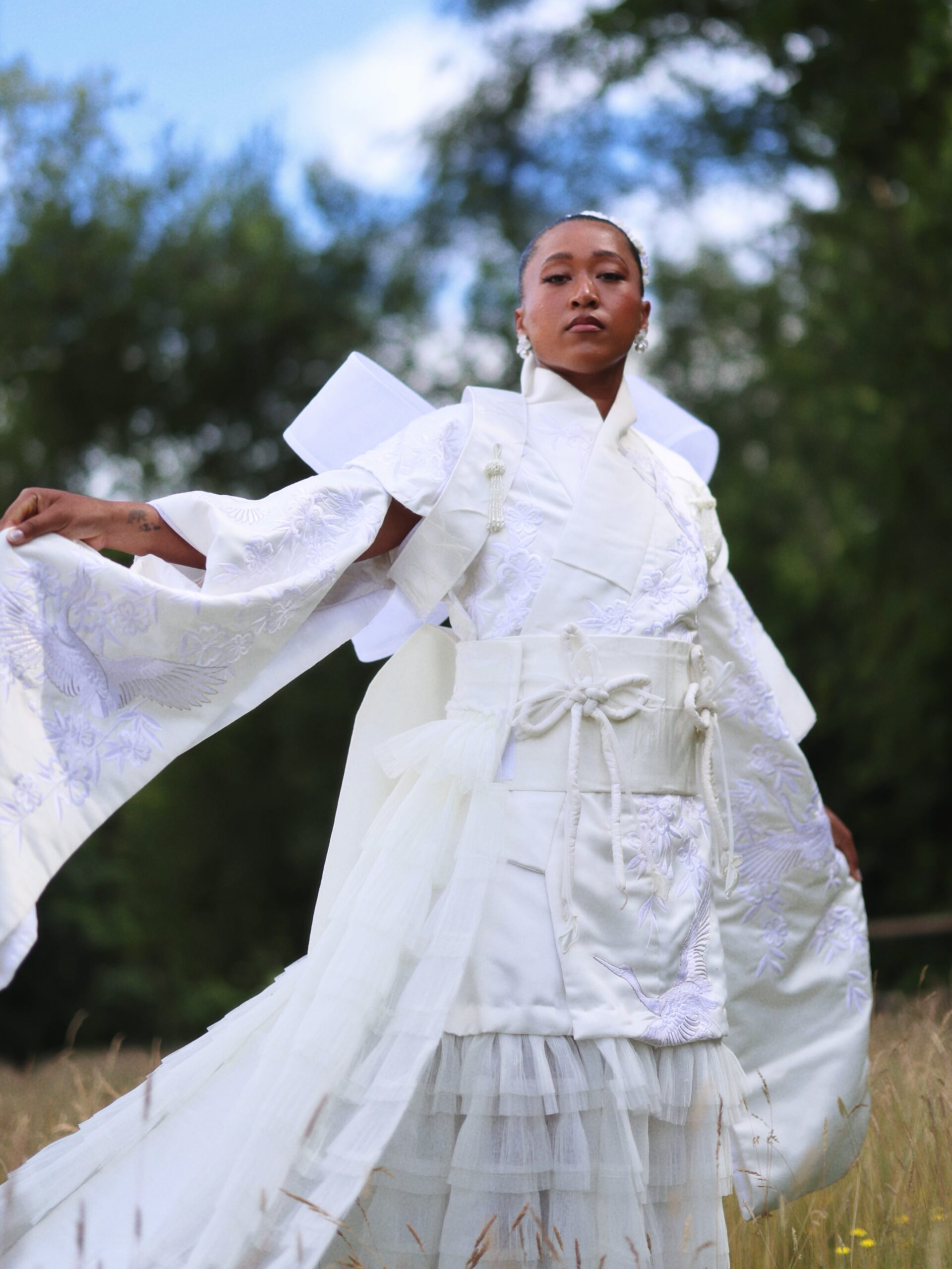 Naomi Osaka holding out the wide sleeve of her embroidered white gown during an outdoor fashion shoot.