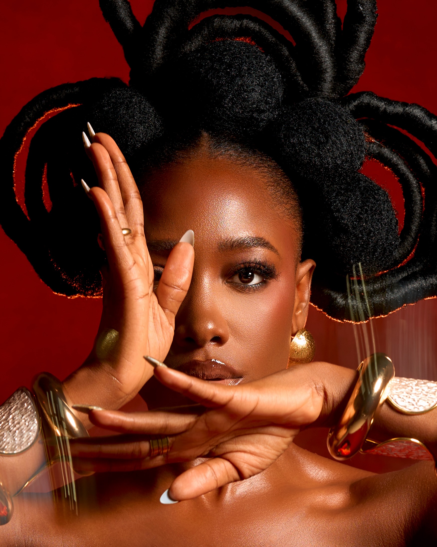 Gbubemi Ejeye framing her face with golden bangles to highlight her minimalist neutral makeup look and textured afro puff updo hairstyle against a deep red studio backdrop.
