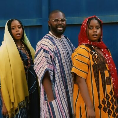 Falz wearing a striped dashiki alongside musical duo Oiza x Meyi in traditional Northern attire standing behind jerrycans in the Na Je music video.