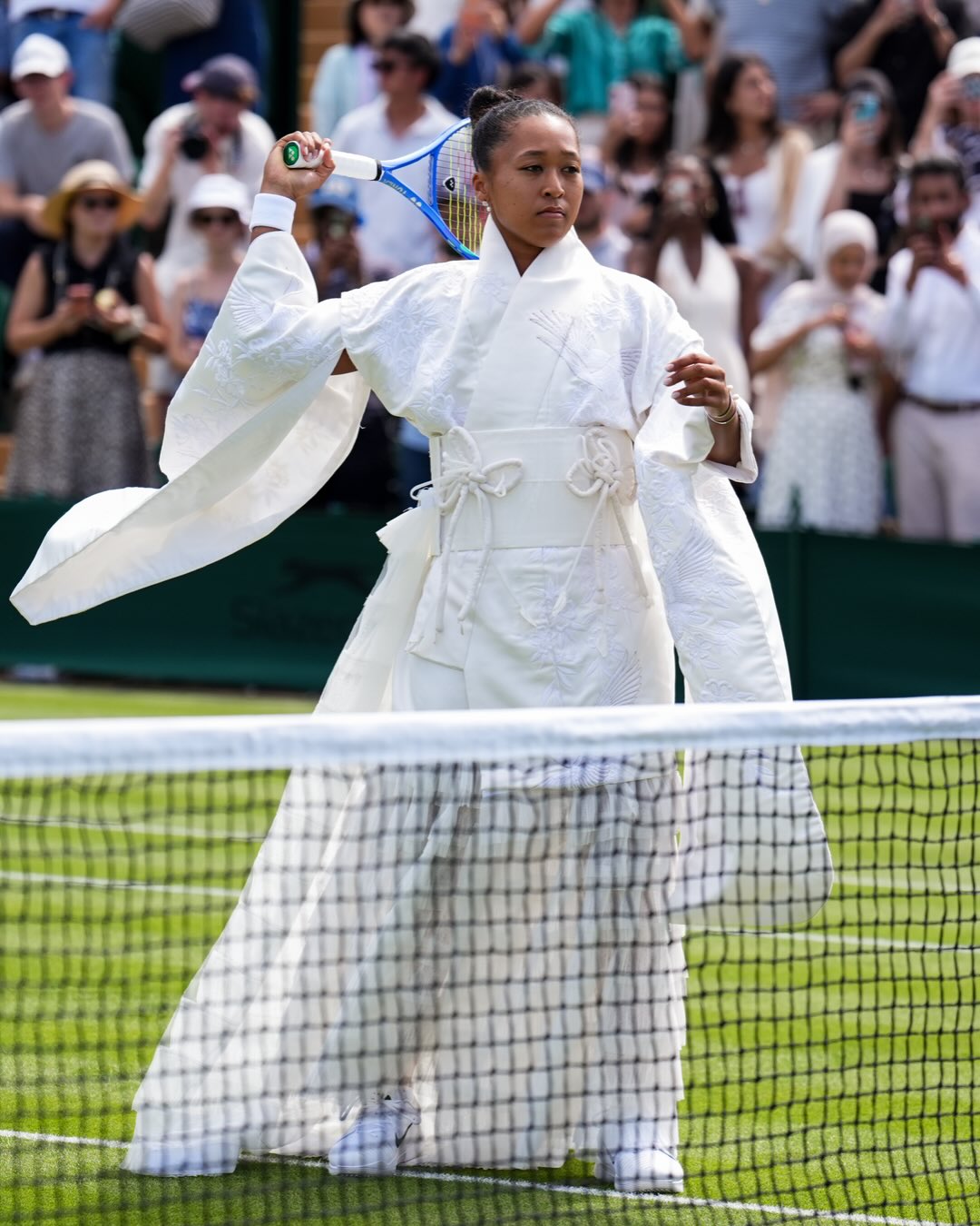 Naomi Osaka standing on a grass tennis court in an embroidered white kimono gown with wide sleeves.