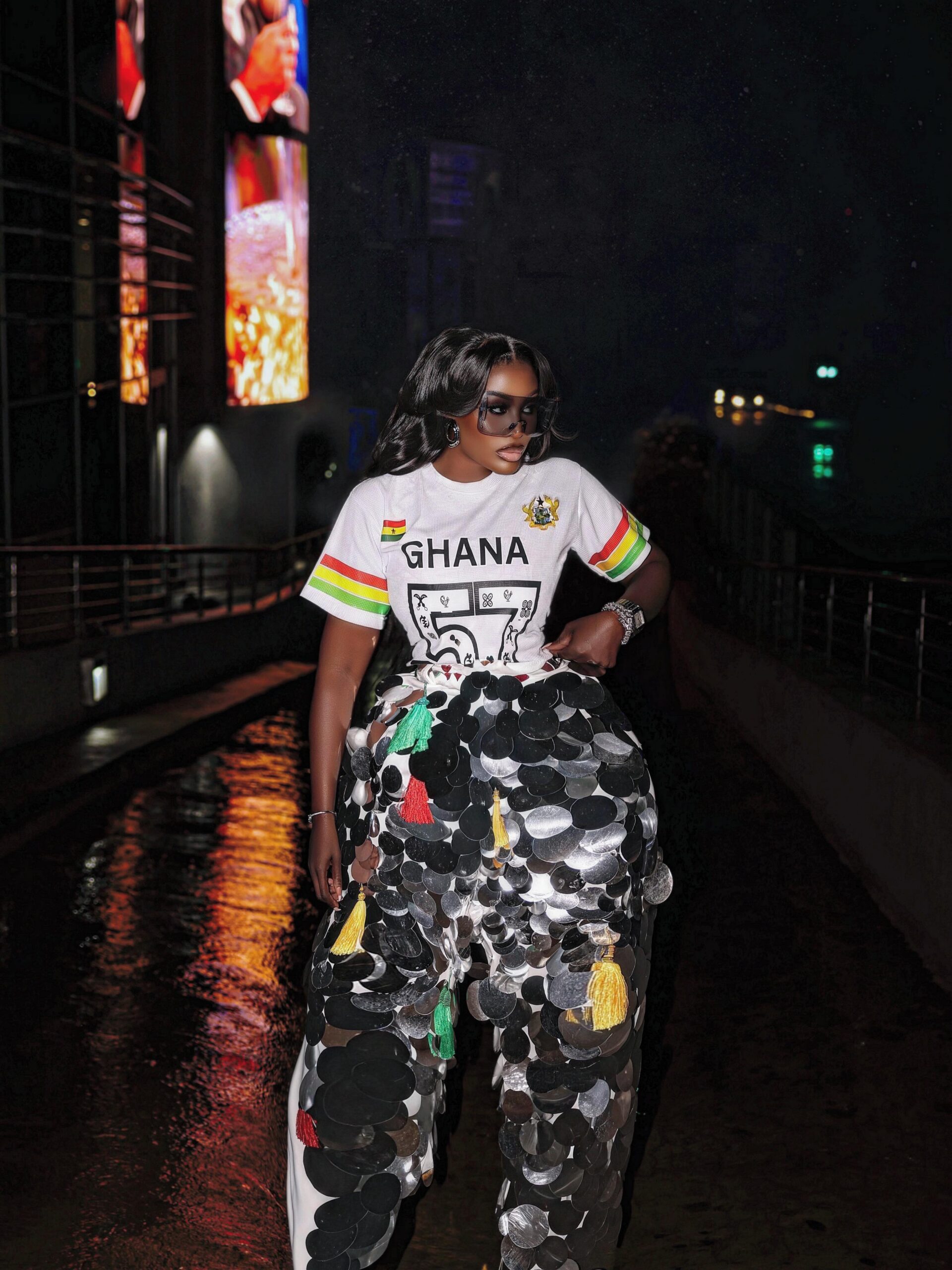 Nana Akua Addo wearing a white custom jersey tee reading GHANA 57 paired with wide-leg trousers covered in silver metallic discs and tri-colour yarn tassels.