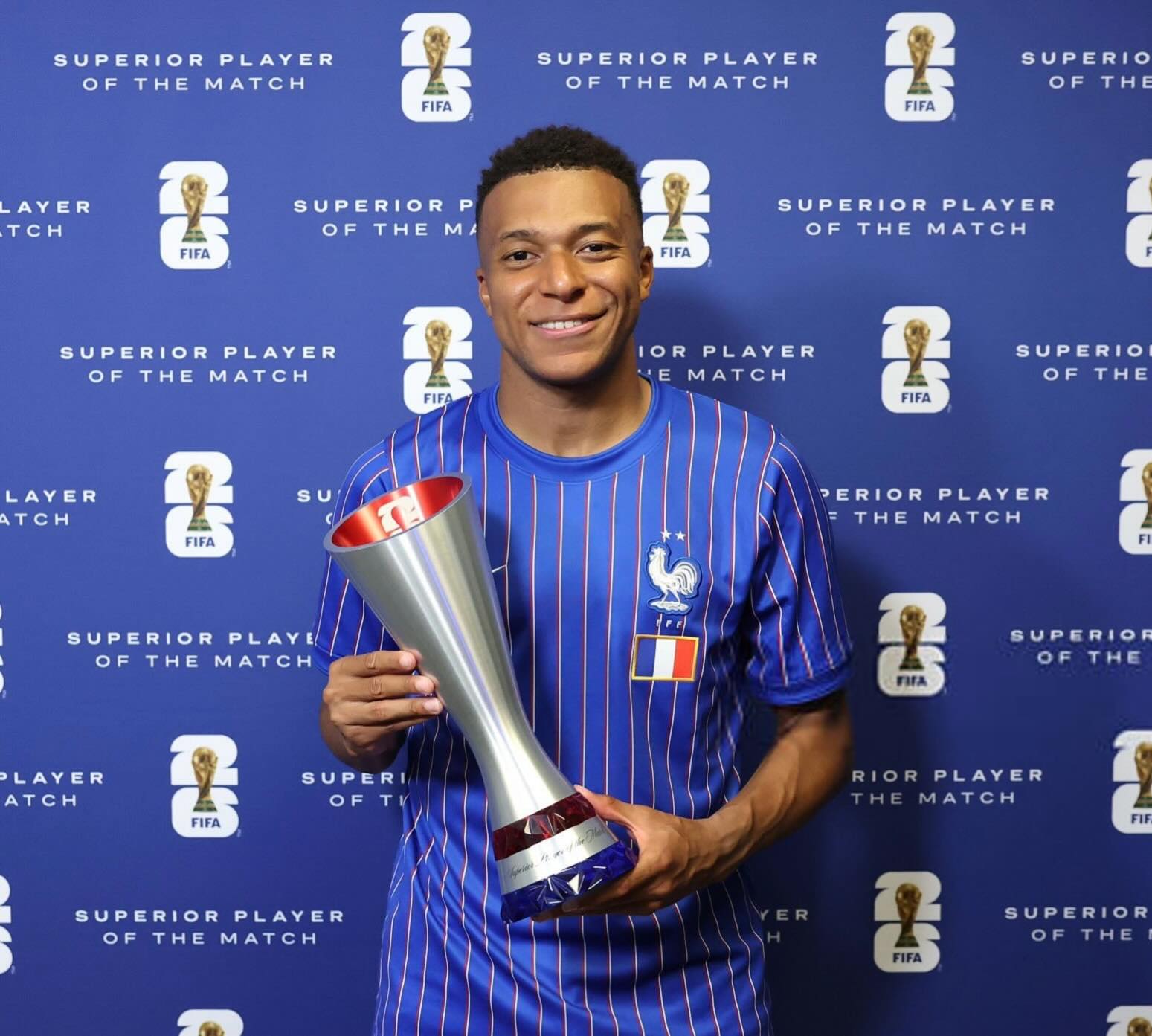 Kylian Mbappe of Cameroonian and Algerian descent smiling while holding a silver FIFA Player of the Match trophy against a blue branded backdrop.