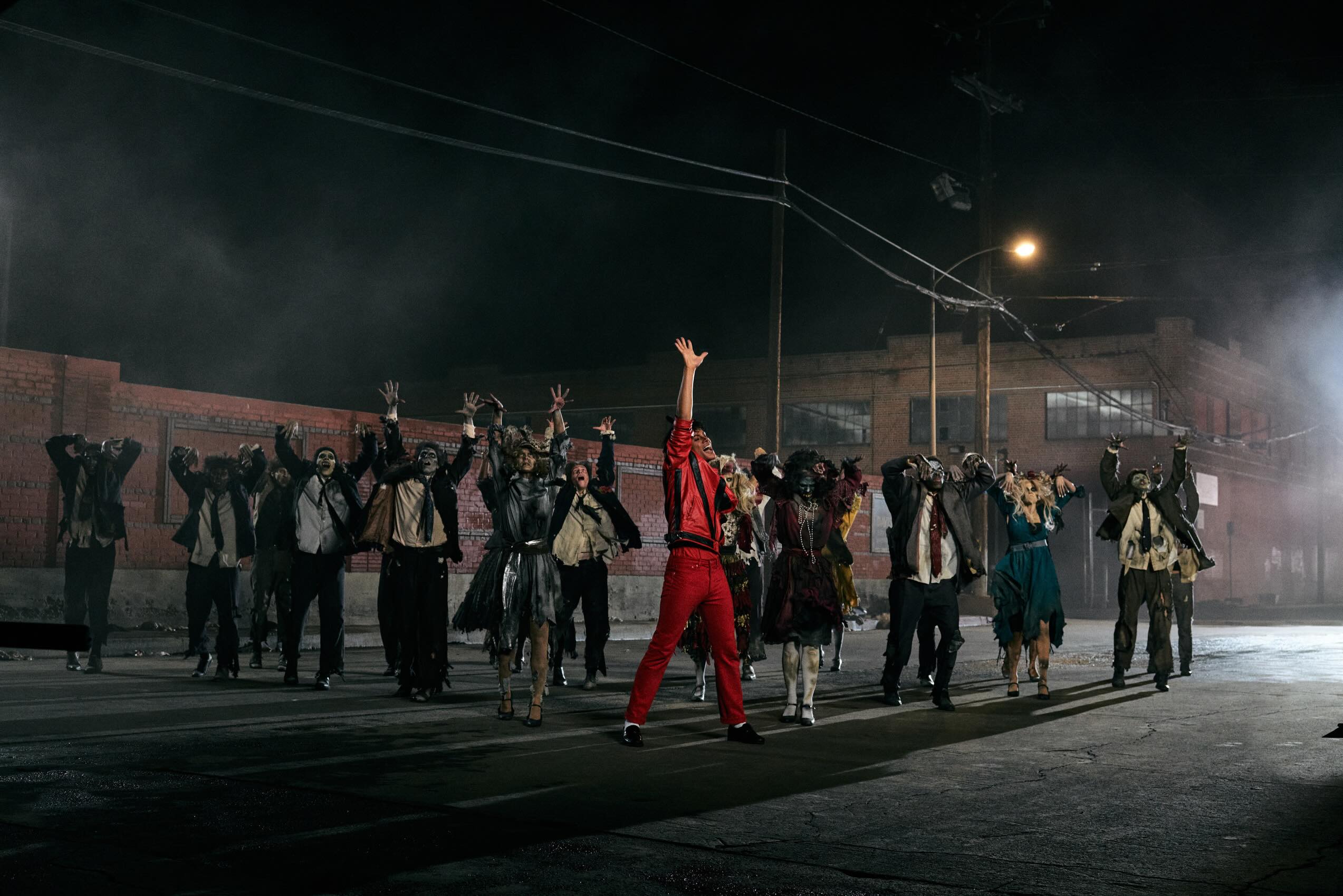 Jaafar Jackson in a red leather jacket and matching red trousers performing the iconic Thriller dance routine on a dark street set surrounded by backing dancers dressed as zombies.
