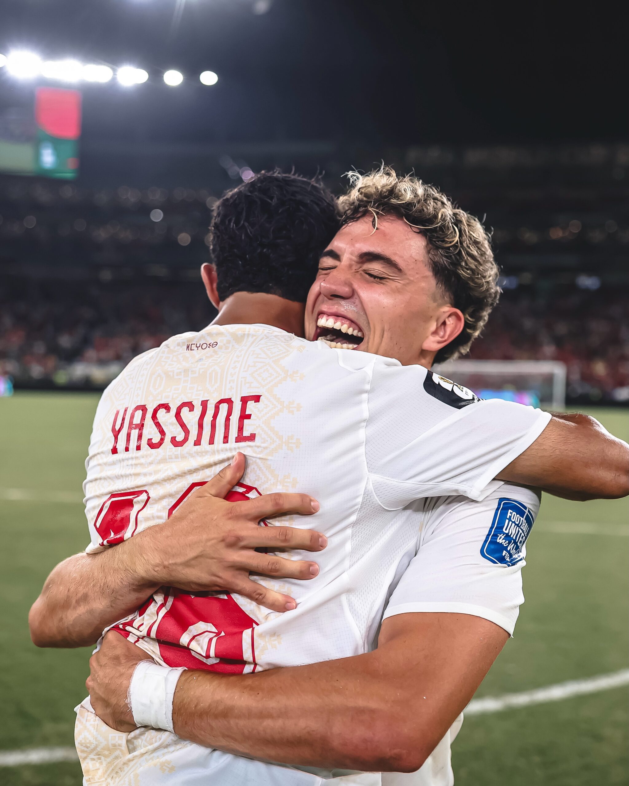 Neil El Aynaoui smiling and hugging goalkeeper Yassine Bounou from behind on the pitch to celebrate knockout qualification.