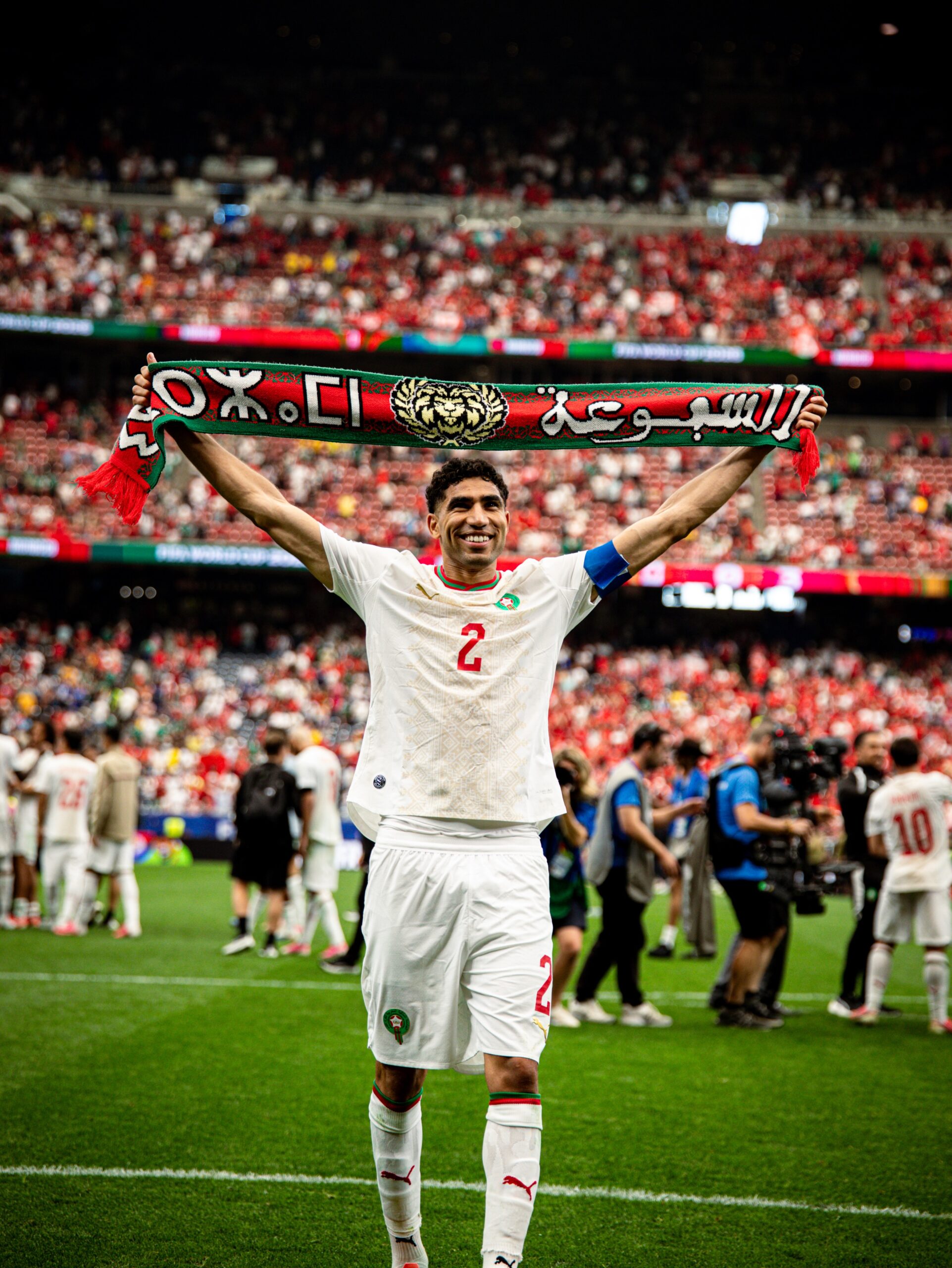 Moroccan defender Achraf Hakimi smiling on the pitch and holding up a green and red supporters' scarf in front of packed stadium stands.
