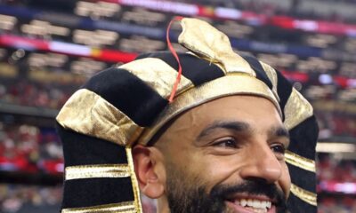 Mohamed Salah smiling on the football field while wearing a black and gold Egyptian pharaoh headdress.