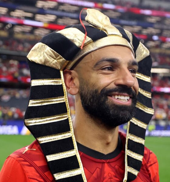 Mohamed Salah smiling on the football field while wearing a black and gold Egyptian pharaoh headdress.