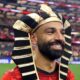 Mohamed Salah smiling on the football field while wearing a black and gold Egyptian pharaoh headdress.