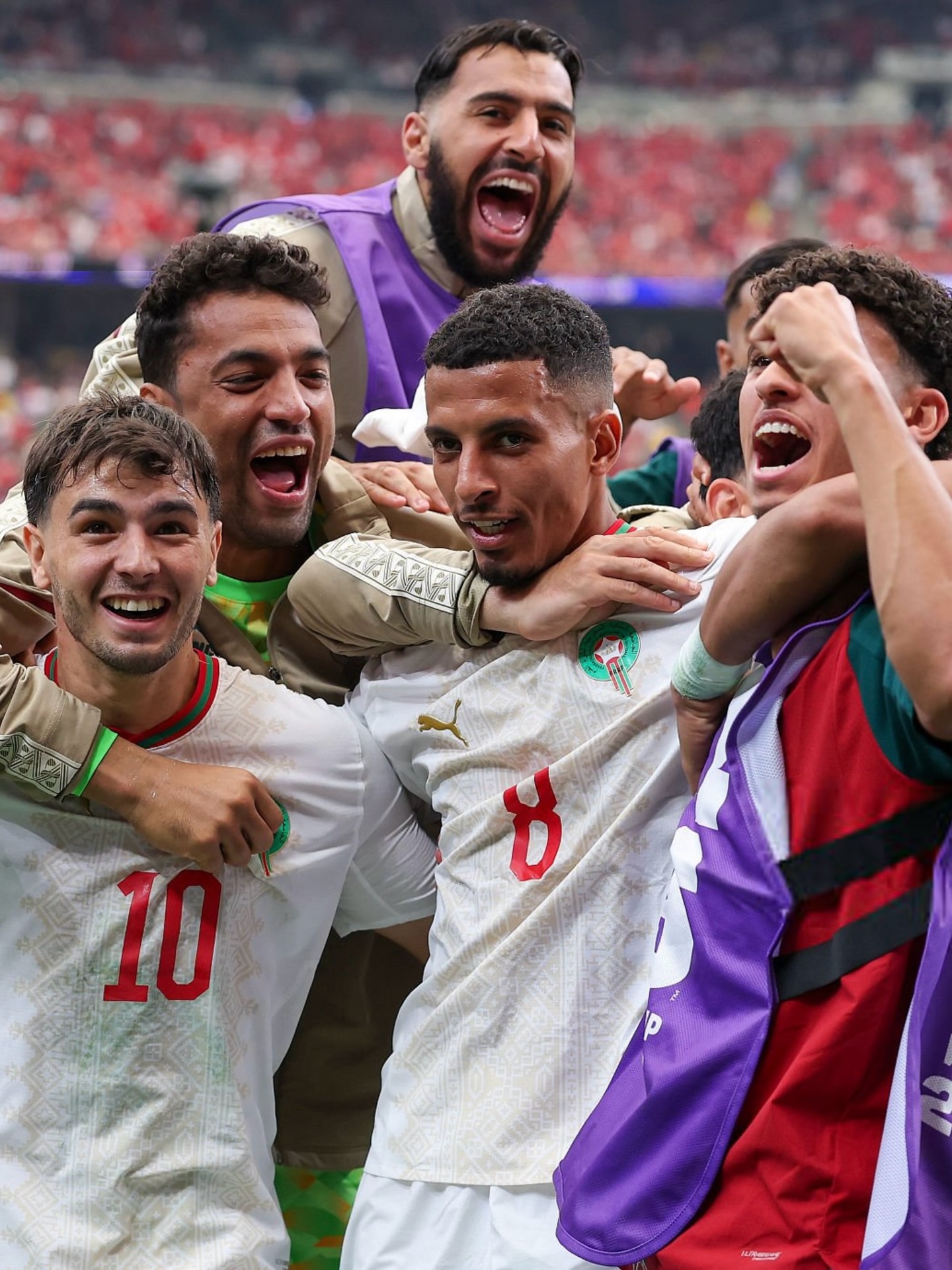 Moroccan midfielder Azzedine Ounahi wearing the number 8 jersey celebrating on the pitch while embraced by teammate Anass Zaroury and squad members.