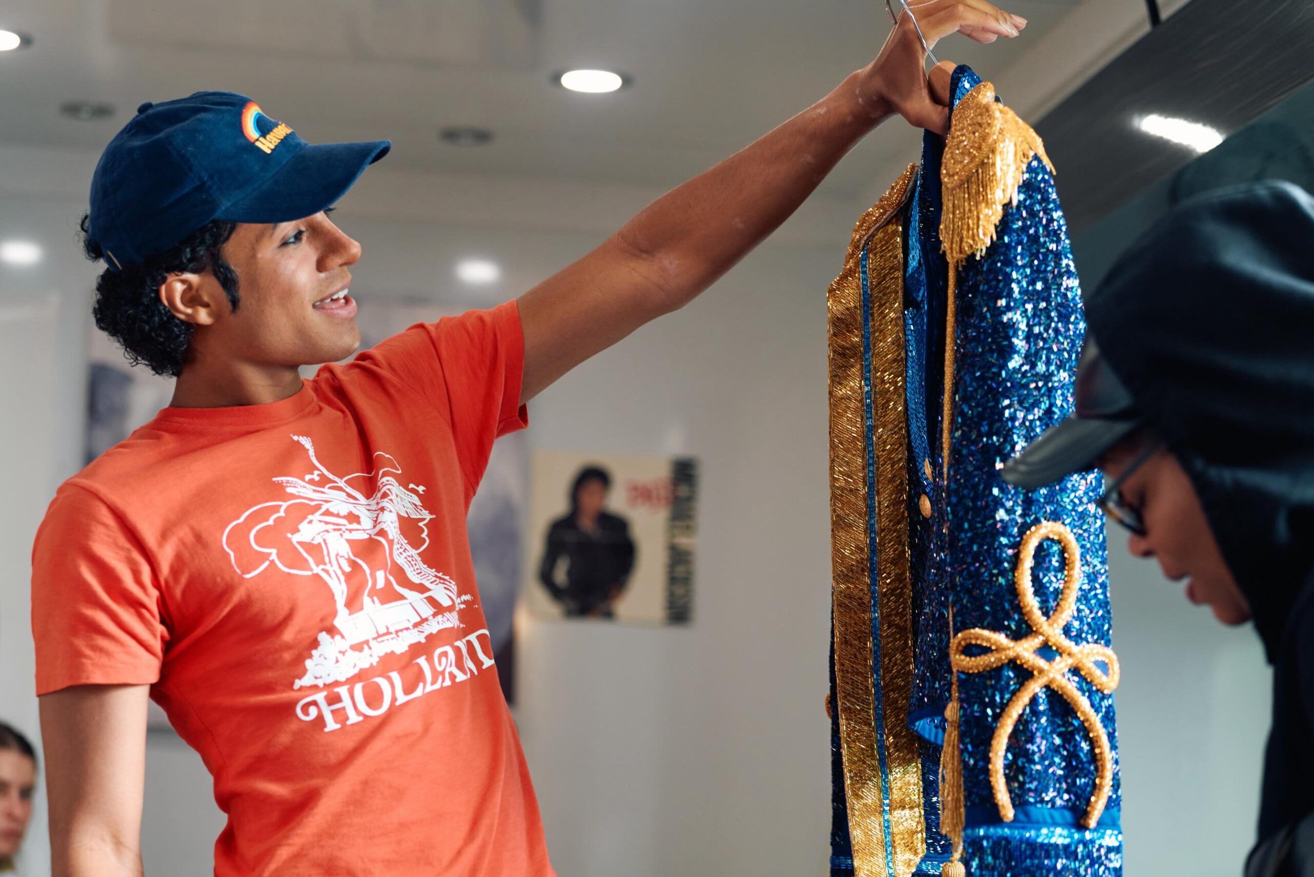Behind-the-scenes photograph of Jaafar Jackson wearing a blue cap and an orange t-shirt, holding up a blue and gold sequined military jacket on a hanger.