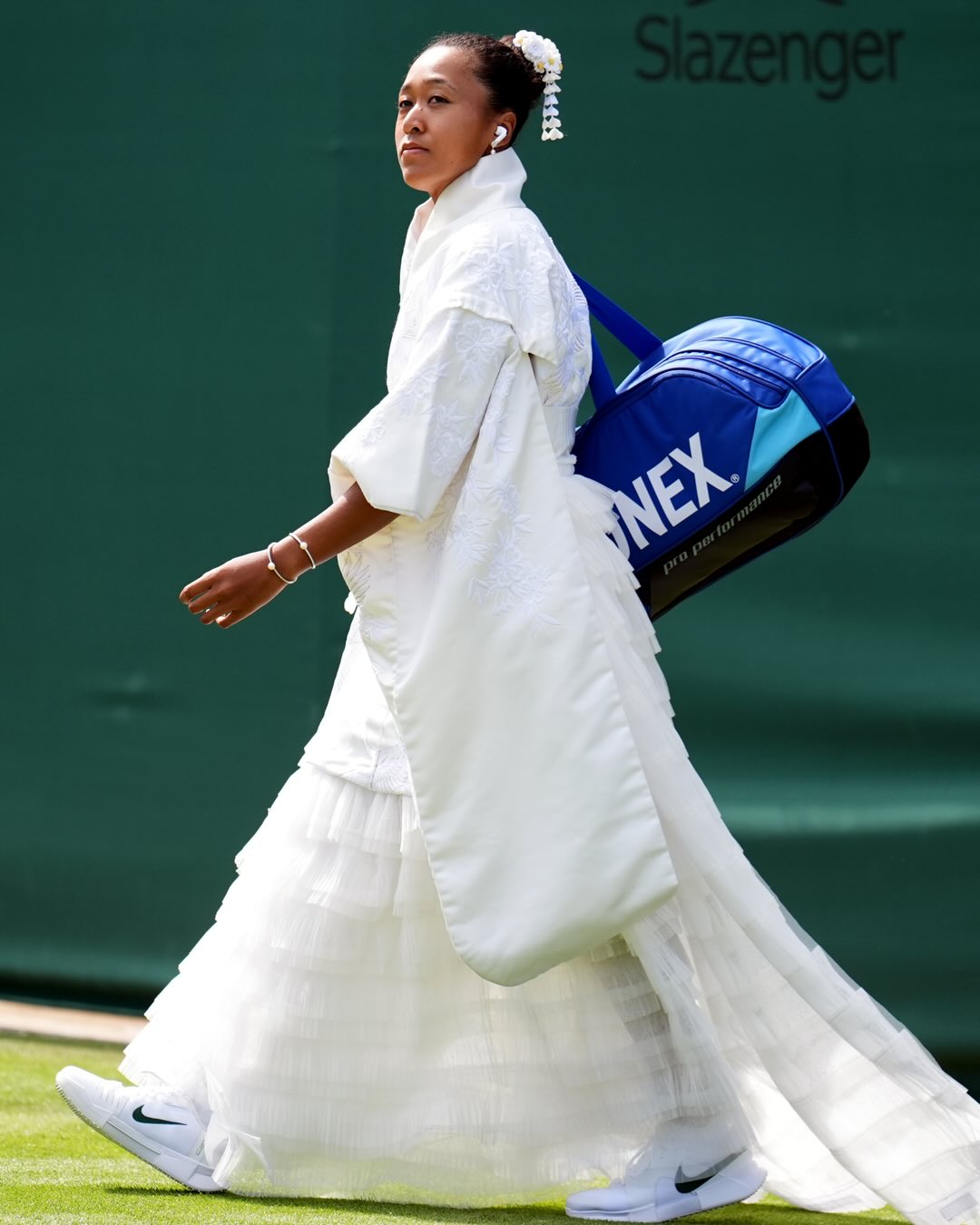 Naomi Osaka walking on grass wearing a white layered tulle skirt, white Nike trainers, and carrying a blue Yonex bag.
