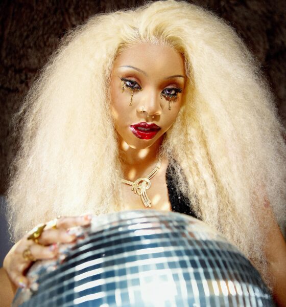 ave with voluminous blonde hair, teardrop graphic eye makeup, a gold necklace, and her hands resting on a reflective disco ball.