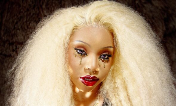 ave with voluminous blonde hair, teardrop graphic eye makeup, a gold necklace, and her hands resting on a reflective disco ball.