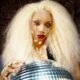 ave with voluminous blonde hair, teardrop graphic eye makeup, a gold necklace, and her hands resting on a reflective disco ball.