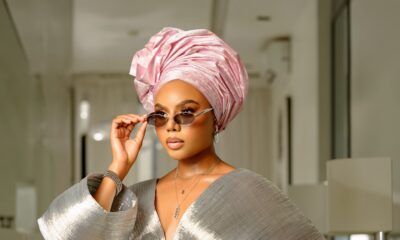 Nancy Isime adjusting silver-framed oval sunglasses while wearing a metallic silver pleated top with dramatic sleeves, an embroidered corset, and a pink gele.