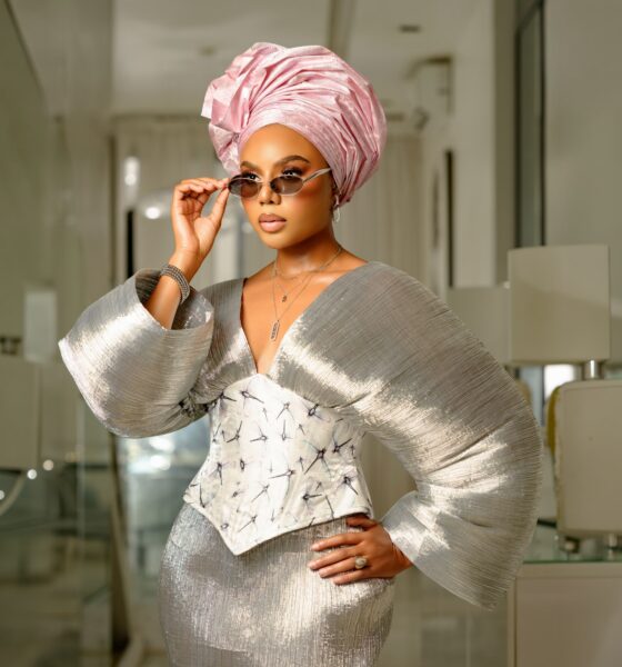 Nancy Isime adjusting silver-framed oval sunglasses while wearing a metallic silver pleated top with dramatic sleeves, an embroidered corset, and a pink gele.