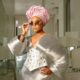 Nancy Isime adjusting silver-framed oval sunglasses while wearing a metallic silver pleated top with dramatic sleeves, an embroidered corset, and a pink gele.
