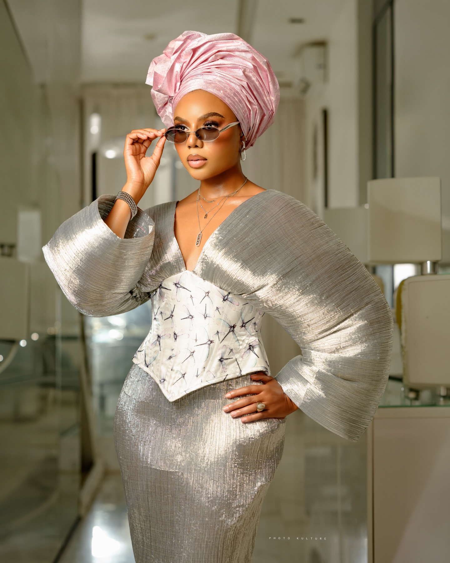 Nancy Isime adjusting silver-framed oval sunglasses while wearing a metallic silver pleated top with dramatic sleeves, an embroidered corset, and a pink gele.