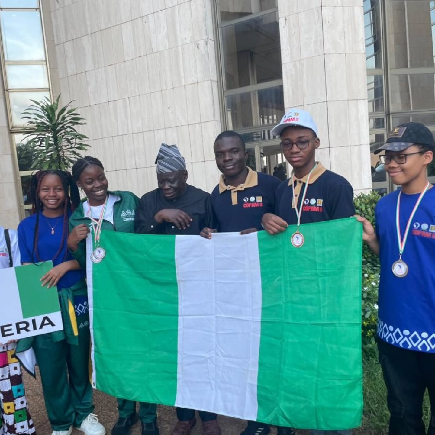 Six Team Nigeria students proudly display their three silver medals and one bronze medal while holding the Nigerian flag outside the Pan-African Mathematics Olympiad venue.
