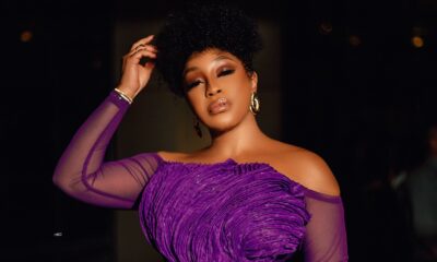 Rita Dominic holding a woven tan leather clutch bag to highlight the intricate textured lines of her off-the-shoulder purple peplum dress bodice.