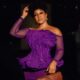 Rita Dominic holding a woven tan leather clutch bag to highlight the intricate textured lines of her off-the-shoulder purple peplum dress bodice.