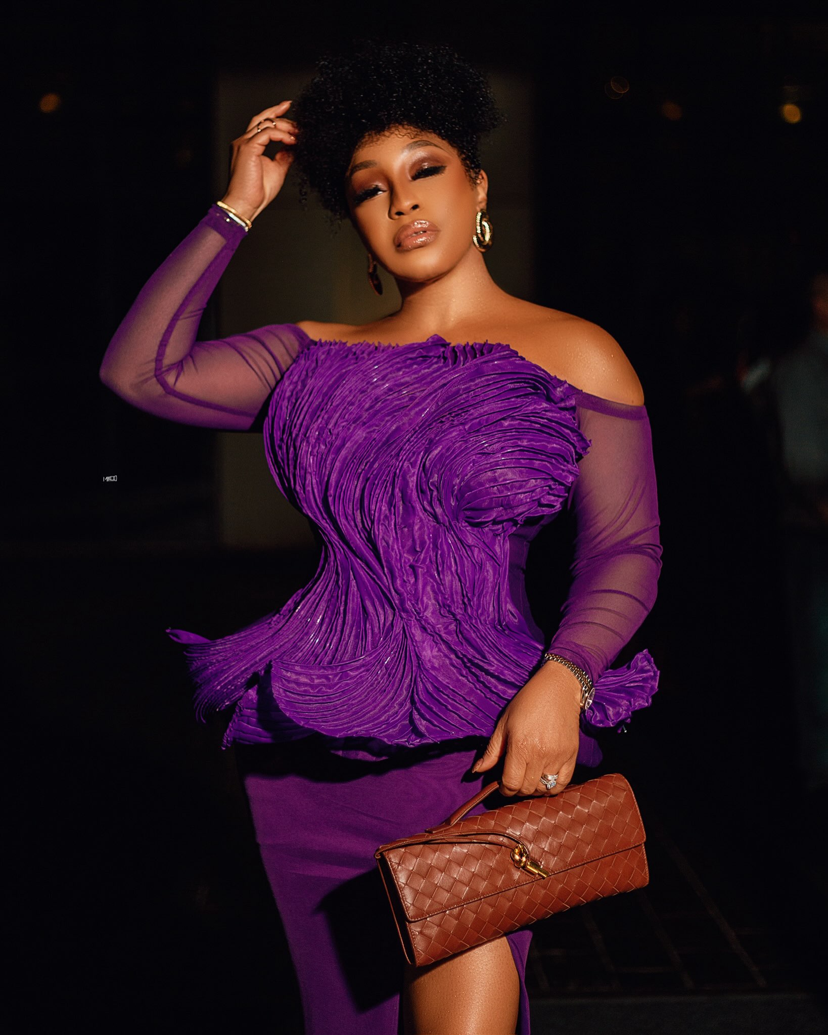 Rita Dominic holding a woven tan leather clutch bag to highlight the intricate textured lines of her off-the-shoulder purple peplum dress bodice.