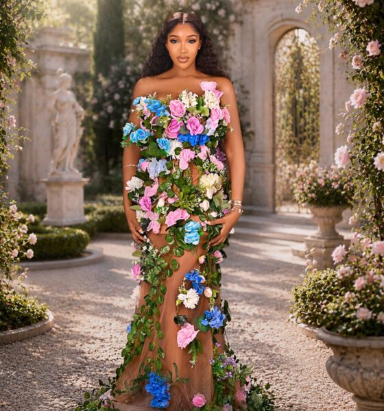Idia Aisien standing in a stone courtyard wearing a strapless flesh-toned gown adorned with trailing green vines and multi-coloured roses.