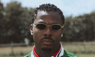 Asake wearing a green knit polo with red floral embroidery, gold oval sunglasses, and gold bracelets in the MCBH music video.