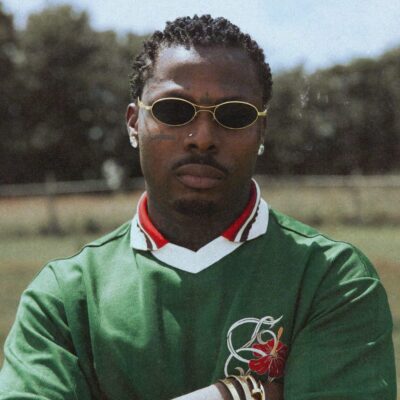 Asake wearing a green knit polo with red floral embroidery, gold oval sunglasses, and gold bracelets in the MCBH music video.