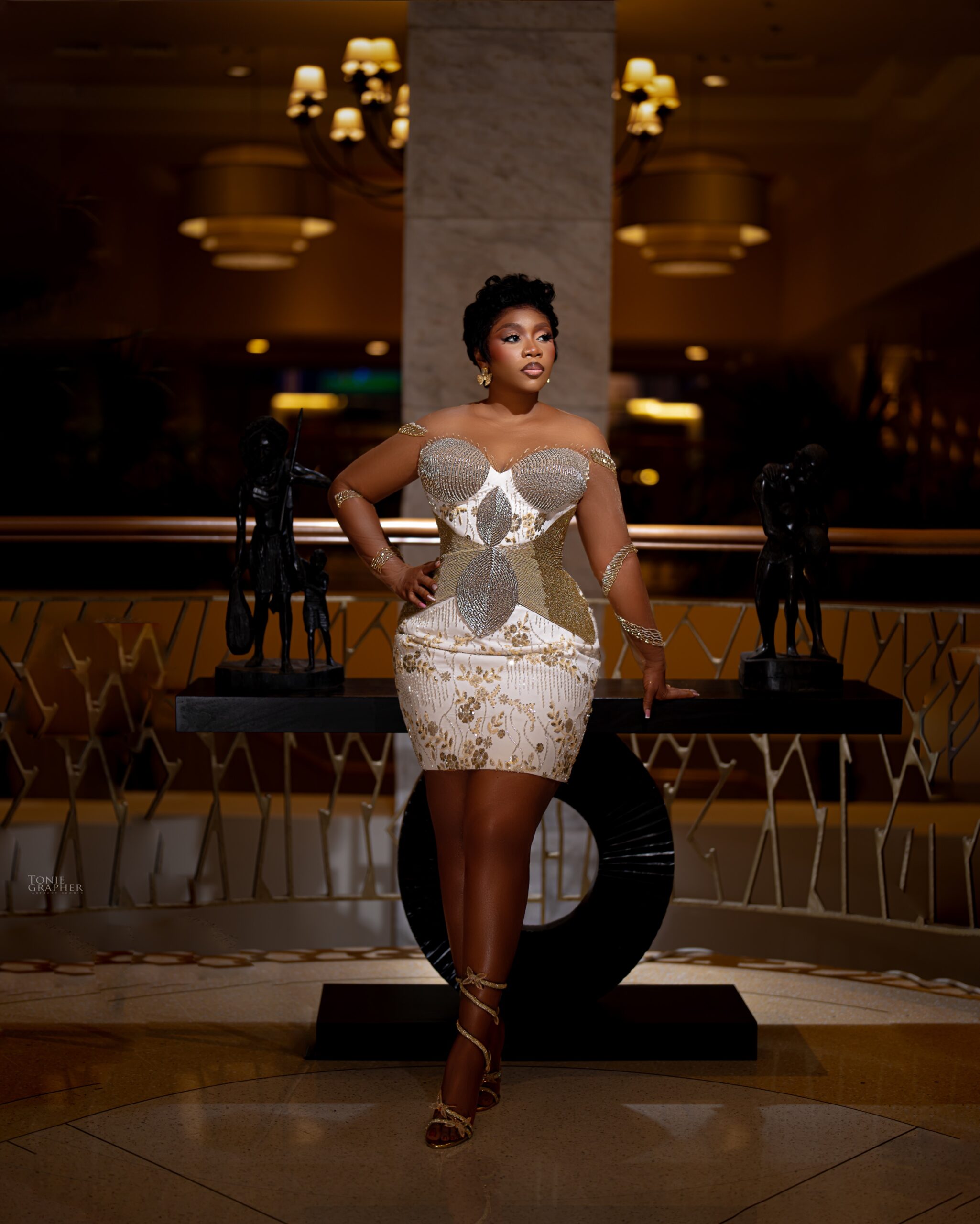 Wumi Toriola posing in a strapless white corset mini dress heavily decorated with silver and gold crystals, abstract leaf-shaped waist panels, and gold filigree embroidery.