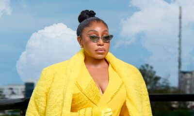 Rita Dominic wearing a plush yellow faux-fur maxi coat draped over a tailored yellow blazer with crystal-embellished sunglasses on a balcony.