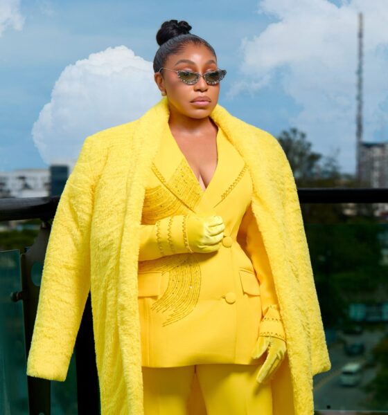 Rita Dominic wearing a plush yellow faux-fur maxi coat draped over a tailored yellow blazer with crystal-embellished sunglasses on a balcony.