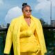 Rita Dominic wearing a plush yellow faux-fur maxi coat draped over a tailored yellow blazer with crystal-embellished sunglasses on a balcony.