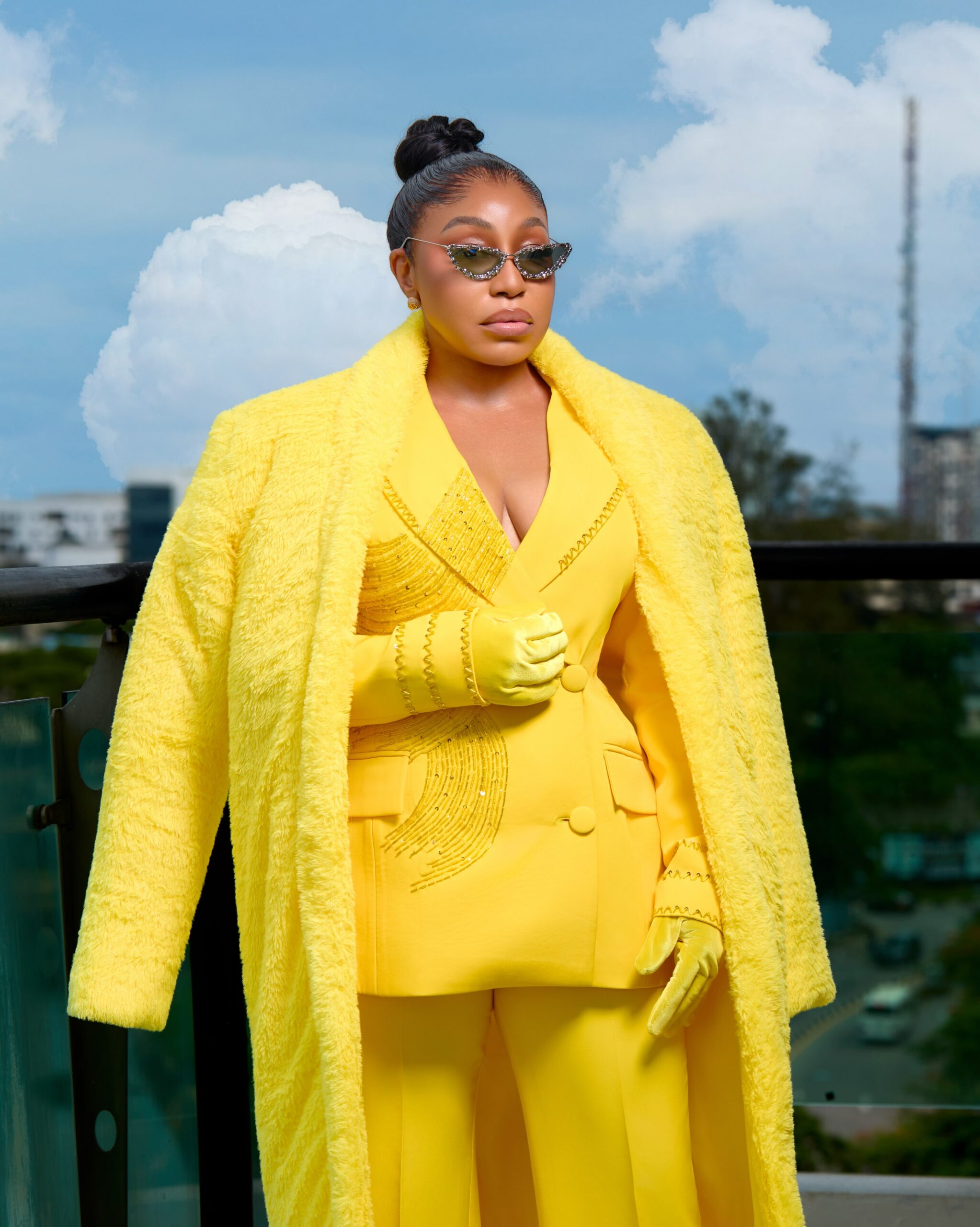 Rita Dominic wearing a plush yellow faux-fur maxi coat draped over a tailored yellow blazer with crystal-embellished sunglasses on a balcony.