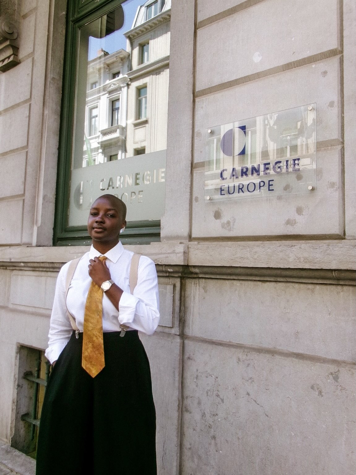 BBNaija star Saskay stands outside Carnegie Europe in Brussels wearing a white shirt, beige suspenders, black high-waisted trousers, and a gold tie.