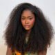 Dede Ashiogwu wears dark brown lip liner with nude-pink lipstick and clear gloss framed by long curly hair.