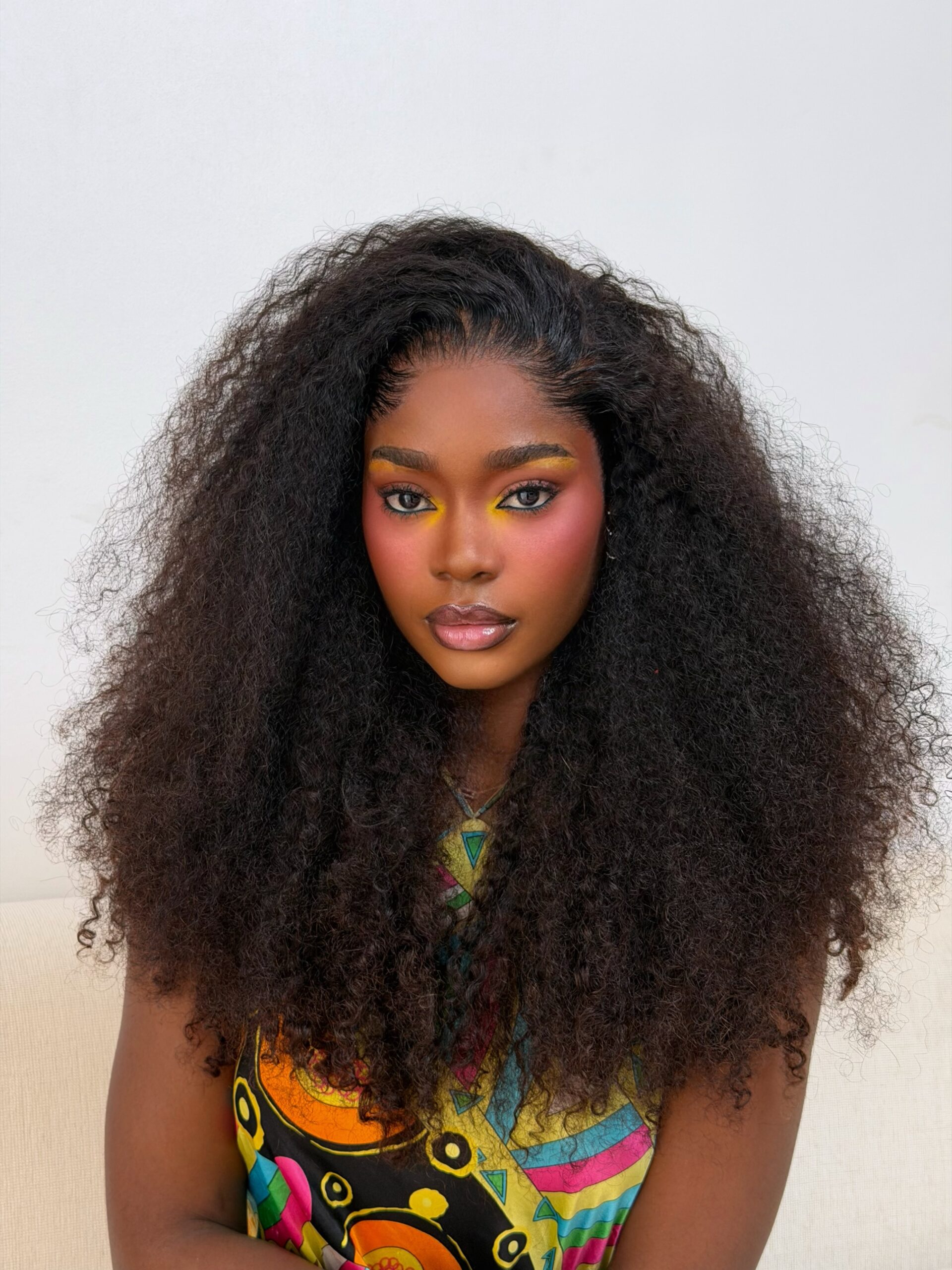 Dede Ashiogwu wears dark brown lip liner with nude-pink lipstick and clear gloss framed by long curly hair.
