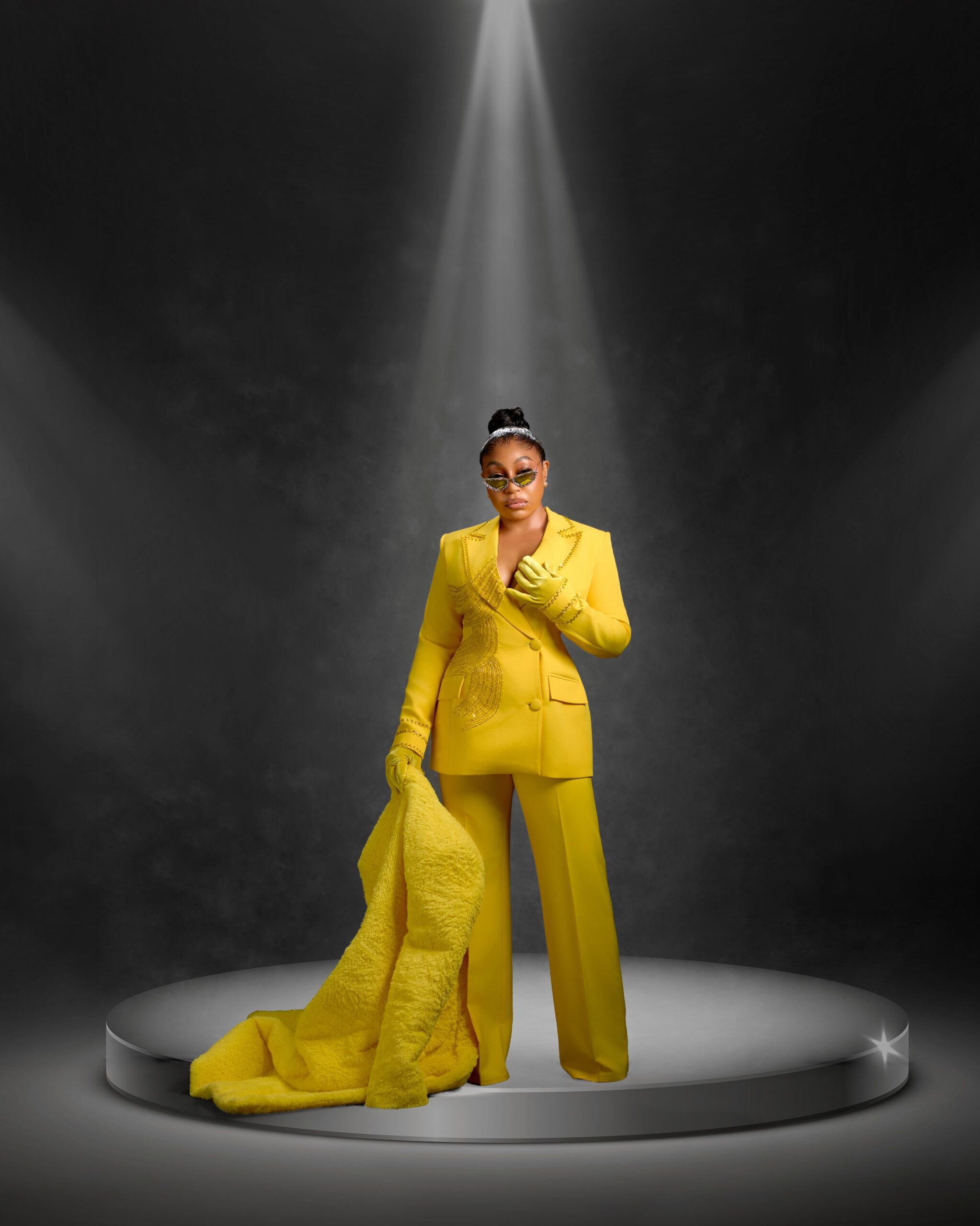 Actress Rita Dominic posing on a circular stage under a spotlight wearing a monochromatic yellow tailored pantsuit and matching satin gloves.