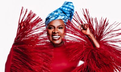 Angelique Kidjo smiling in her crimson-red Imane Ayissi Couture gown with oversized, structured red raffia fringe sleeves and a blue tie-dye headwrap.