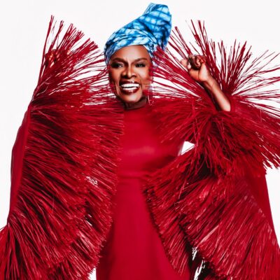 Angelique Kidjo smiling in her crimson-red Imane Ayissi Couture gown with oversized, structured red raffia fringe sleeves and a blue tie-dye headwrap.