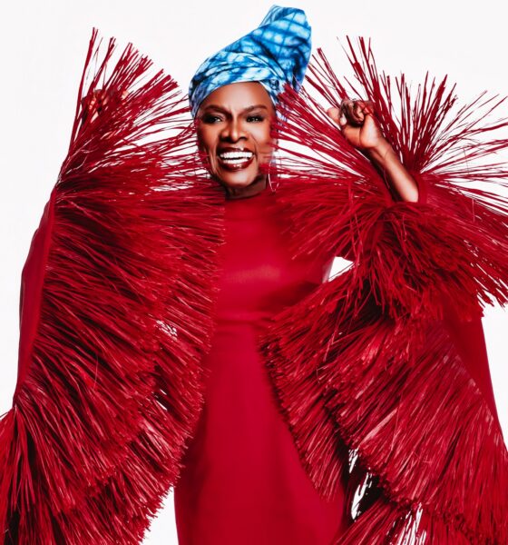 Angelique Kidjo smiling in her crimson-red Imane Ayissi Couture gown with oversized, structured red raffia fringe sleeves and a blue tie-dye headwrap.