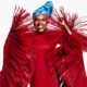 Angelique Kidjo smiling in her crimson-red Imane Ayissi Couture gown with oversized, structured red raffia fringe sleeves and a blue tie-dye headwrap.