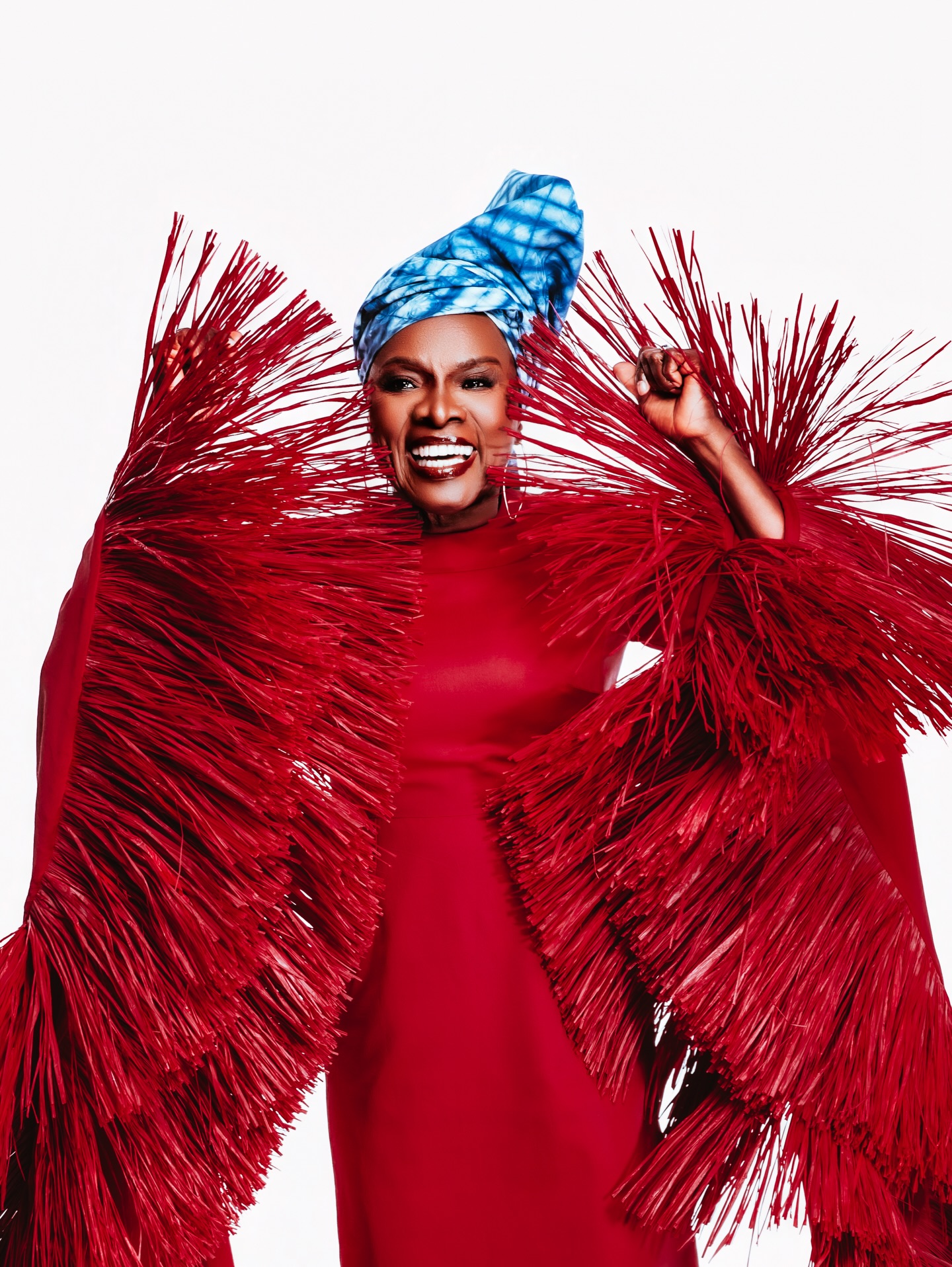 Angelique Kidjo smiling in her crimson-red Imane Ayissi Couture gown with oversized, structured red raffia fringe sleeves and a blue tie-dye headwrap.