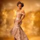 Toke Makinwa in a peach mermaid-cut traditional gown with a gold basket-weave central panel, asymmetric high-low flared hemline, and metallic strappy sandals.