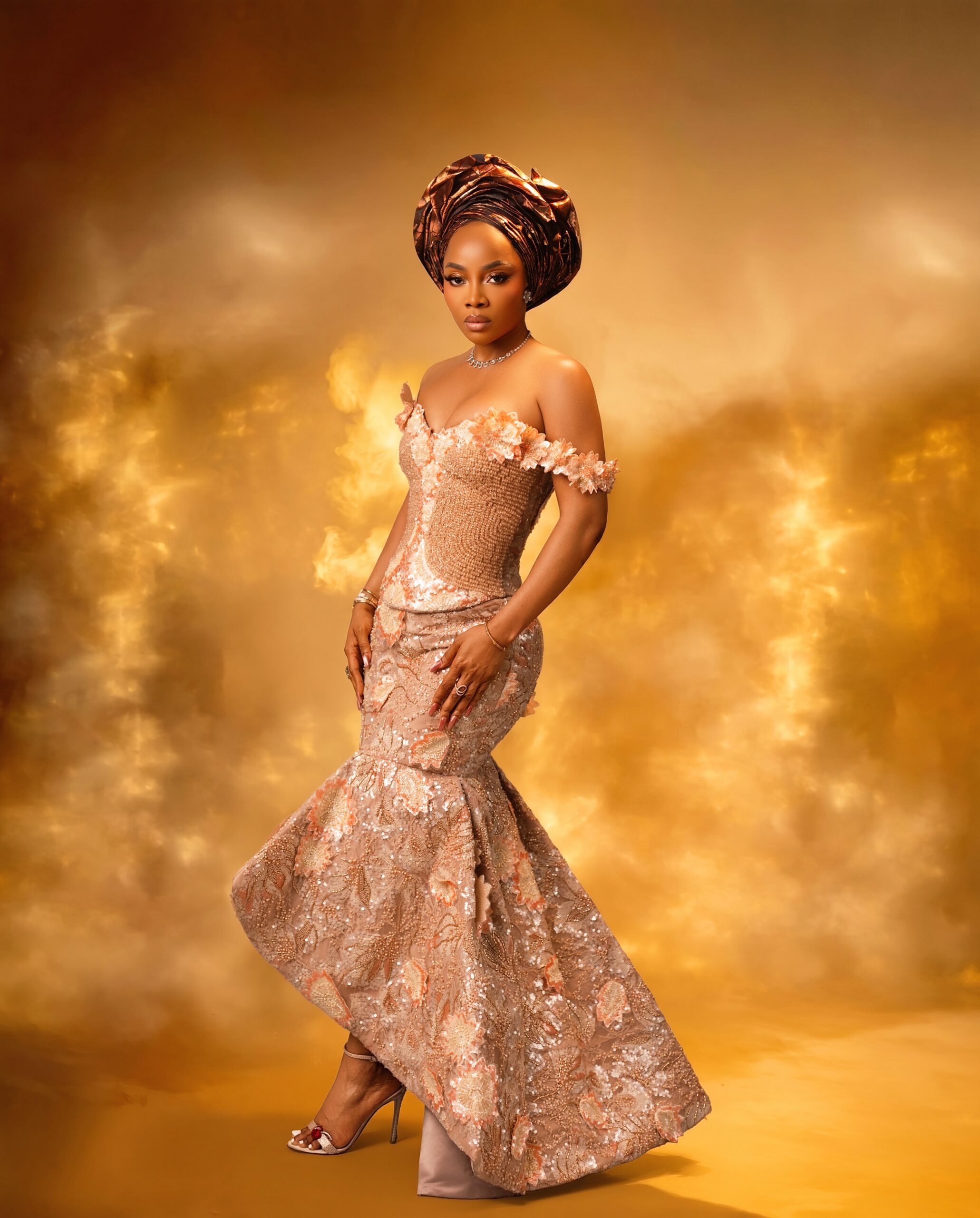 Toke Makinwa in a peach mermaid-cut traditional gown with a gold basket-weave central panel, asymmetric high-low flared hemline, and metallic strappy sandals.