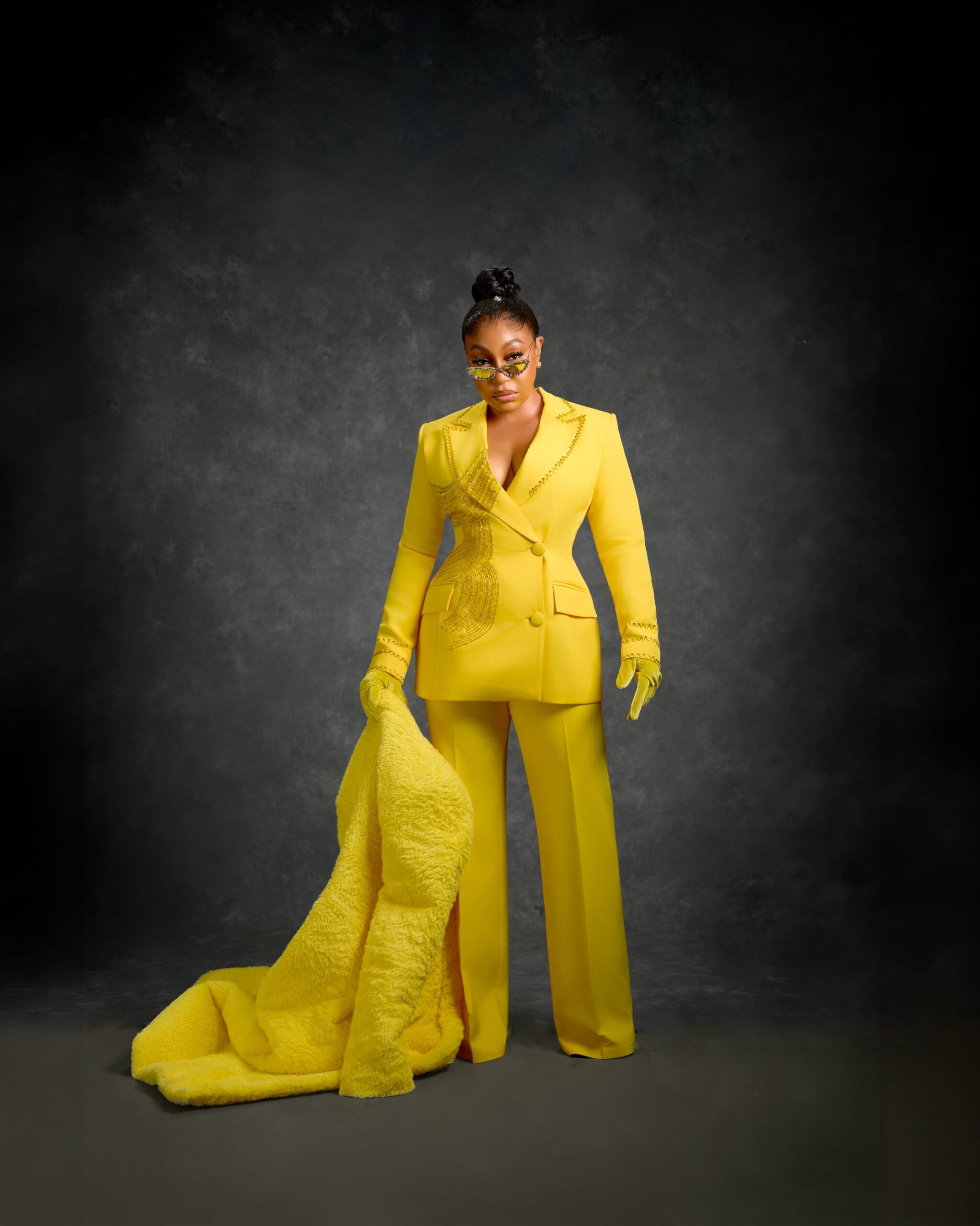 Rita Dominic standing against a textured grey studio backdrop in a yellow structured blazer with linear beadwork and wide-leg trousers.