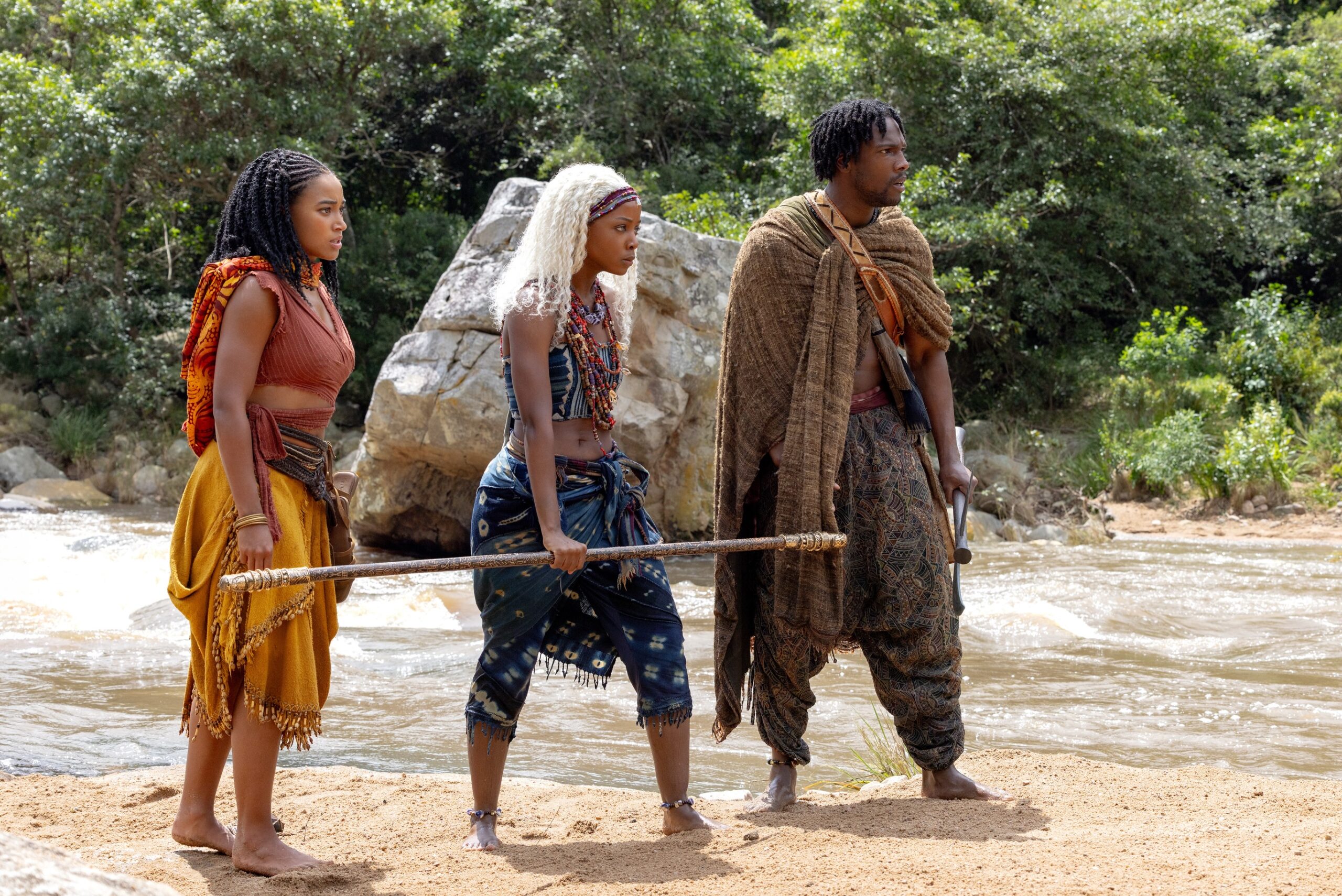 Amandla Stenberg as Princess Amari, Thuso Mbedu as Zélie Adebola, and Tosin Cole as Tzain standing on a sandy riverbank in the kingdom of Orïsha.